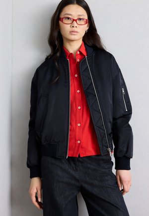 Fiatal nő hosszú sötét hajjal, piros szemüveget, piros gombos inget, fekete bomber dzsekit és sötét nadrágot viselve, világosszürke fal előtt áll.