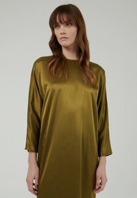 Vestido de satén verde oliva con corte holgado, cuello redondo y mangas tres cuartos. Textura suave con detalles de diseño mínimos.