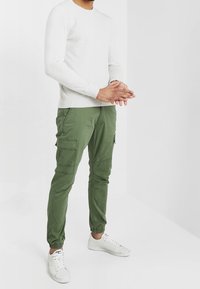 Lichtgrijs long-sleeve shirt gecombineerd met groene cargobroek met meerdere zakken en elastische manchetten, aangevuld met witte sneakers.