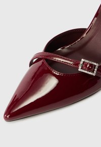 Μπορντό παπούτσι υψηλού τακουνιού από πατενέ δέρμα με μυτερή μύτη και διακοσμητική λωρίδα που διαθέτει εμβληματική αγκράφα με πέτρες. Λείος υφής.