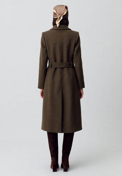 Cappotto lungo in lana marrone con vita a cintura, colletto a scollo a V e spacco centrale sul retro. Abbinato a stivali al ginocchio in suede marrone scuro e a una sciarpa in seta.