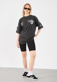 T-shirt oversized preta de algodão com estampas gráficas, combinada com calções de bicicleta pretos e ténis pretos com riscas brancas e meias até ao tornozelo.