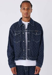 Chaqueta de mezclilla en azul oscuro con patrón de pespunte blanco. Cuenta con dos bolsillos en el pecho y herrajes plateados, con un cuello clásico y cierre de botones.
