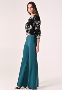 Zwart top met bloemenpatroon en driekwart mouwen, gecombineerd met high-waisted tealovertrekken en zwarte blokhak sandalen.