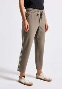 Persoon in taupe joggerpants met witte en zwarte zijkanten en witte sneakers op een witte achtergrond.