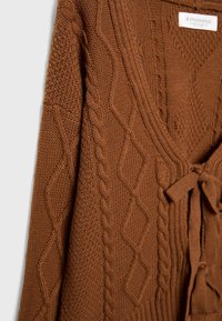 Cardigan marron en maille avec motifs torsadés et losanges, fermeture à nouer à l'avant, et étiquette visible de la marque Stradivarius à l'intérieur du col.