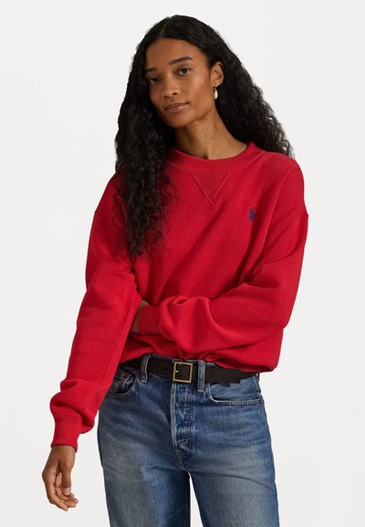 Kvinde med langt krøllet mørkt hår iført rød sweatshirt og blå jeans med sort bælte, stående med armene korset mod en hvid baggrund.