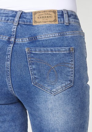 Jean en denim bleu mi-hauteur avec une étiquette de marque beige, un détail de couture visible sur les poches arrière et une texture légèrement usée.