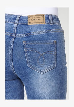 Keskmise kõrgusega sinised denim püksid, millel on beež brändi silt, nähtavad tagakoti õmbluse detailid ja veidi kulunud tekstuur.