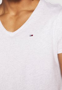 Närbild på en person som bär en vit V-ringad t-shirt med en liten rektangulär röd, vit och blå logotyp på bröstet.
