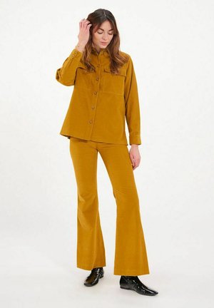 Chemise en velours côtelé moutarde avec un pantalon évasé assorti. La chemise possède une poche avant et une fermeture à boutons. Des bottes noires brillantes complètent la tenue.