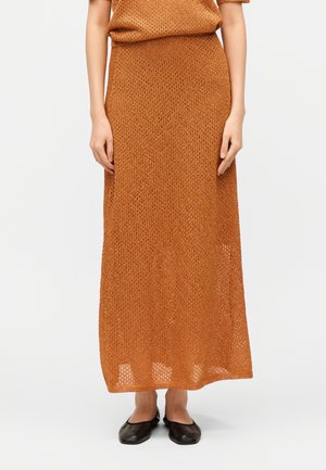 Γυναίκα που φοράει μια μπεζ-πορτοκαλί υφαντή μεγάλη φούστα και αντίστοιχο πάνω μέρος, συνδυασμένα με μαύρα επίπεδα παπούτσια, στέκεται μπροστά σε ένα λευκό φόντο.