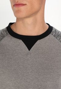 Szary sweatshirt z czarnym prążkowanym dekoltem i teksturowanymi akcentami na ramionach. Posiada V-kształtne przeszycie na klatce piersiowej oraz gładką tkaninę.