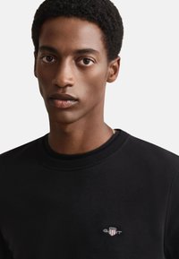 Felpa nera con scollo rotondo, con piccolo logo ricamato sul petto. Il tessuto appare liscio con una silhouette aderente.