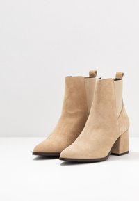 Bottines en suède beige avec un talon bloc, des panneaux latéraux élastiques et un bout arrondi. Texture lisse avec des détails de couture minimaux.