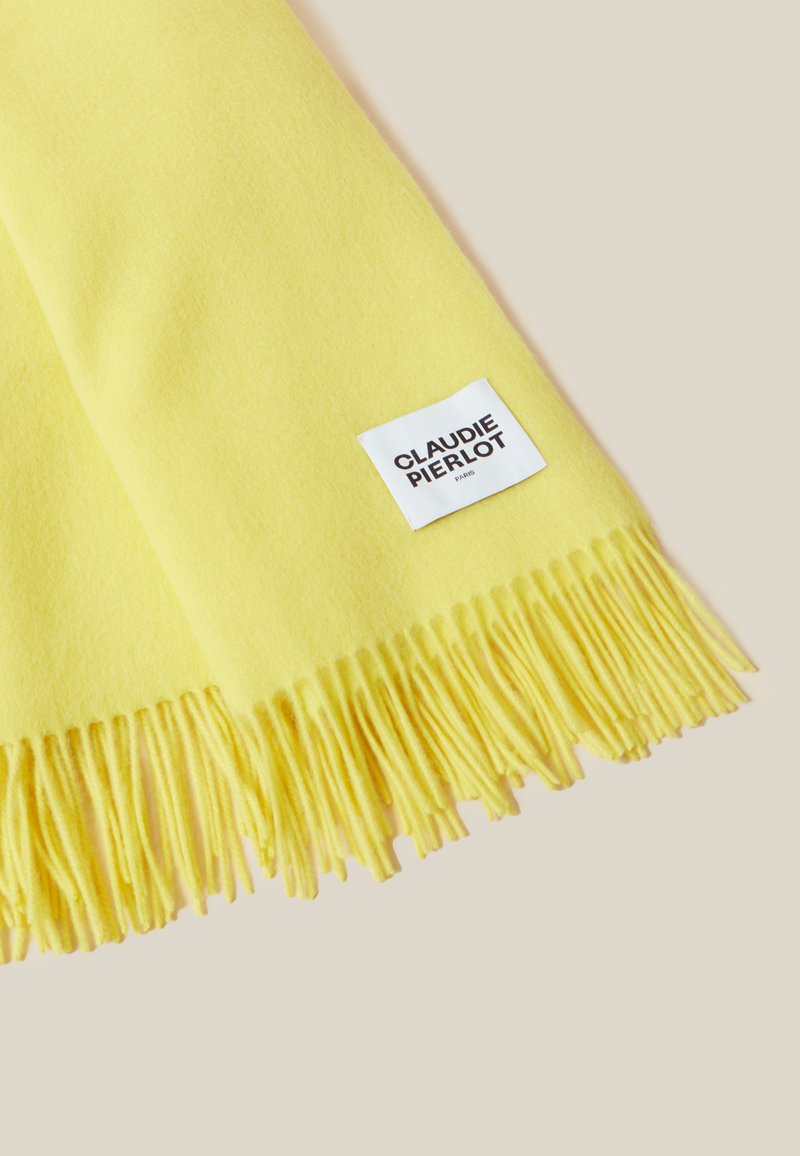 Weicher gelber Schal mit fransigen Kanten und einem weißen Etikett mit der Aufschrift "Claudie Pierlot Paris" auf einem hellbeigen Hintergrund.