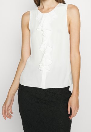 Vrouw draagt een mouwloze witte blouse met ruchtdetail aan de voorkant en een zwarte kanten rok tegen een effen achtergrond.