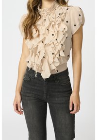 Beige blouse met zwarte stippen, volants aan de voorkant en een hoge volantskraag. G gedragen met donkere jeans, wat een getailleerde silhouet laat zien.