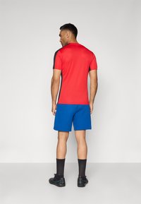 Camisa atlética vermelha de manga curta com detalhes pretos, emparelhada com calções azuis. O conjunto tem um design elegante e é feito de um tecido leve.