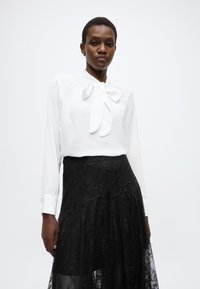 Λευκό πουκάμισο με φιόγκο στη λαιμόκοψη, σε συνδυασμό με μαύρη φούστα από δαντέλα με πολλαπλές υφές και ρέον σιλουέτα.