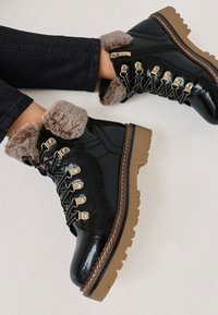 Fekete lakkbőr bokacsizma gyapjúbéléses gallérral, arany színű fémrészletekkel és vastag barna talppal, fűzős és oldalsó cipzárral.