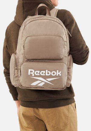 Mochila beige de Reebok con múltiples compartimentos con cremallera, llevada por una persona con sudadera con capucha marrón y pantalones color canela, vista desde atrás.