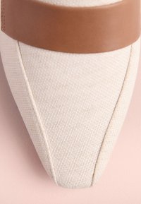 Chaussure en tissu beige à bout pointu, avec une lanière en cuir marron traversant le milieu. Surface texturée et détails de couture visibles.