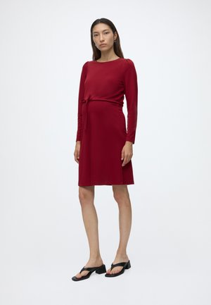 Femme portant une robe rouge mi-longue à manches longues avec une ceinture à la taille, debout, chaussant des sandales noires à talons blocs ouverts sur un fond blanc uni.
