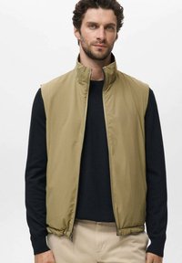 Lichte, mouwloze vest in khaki, met een hoge kraag, rits sluiting en een gladde textuur, gecombineerd met een zwart langärmige shirt.