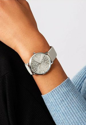 Silberne Analog-Uhr mit einem hellgrauen Silikonarmband, ausgestattet mit einem strukturierten Zifferblatt und großen Ziffern, getragen an einem Handgelenk in einem hellblauen Pullover.