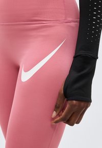 Κοντινό πλάνο ατόμου που φοράει ροζ κολάν Nike με λευκό λογότυπο και μαύρο μακρυμάνικο μπλουζάκι με τρύπα για τον αντίχειρα στο μανίκι.