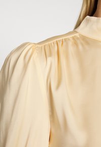 Blusa de cetim amarelo com mangas balão e gola alta. Textura suave, detalhe drapeado na manga e leve brilho.