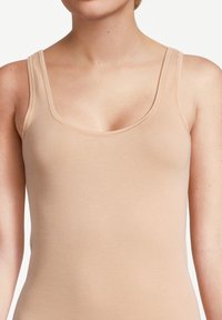 Close-up of a person wearing a sleeveless, beige tank top with double shoulder straps against a plain background.