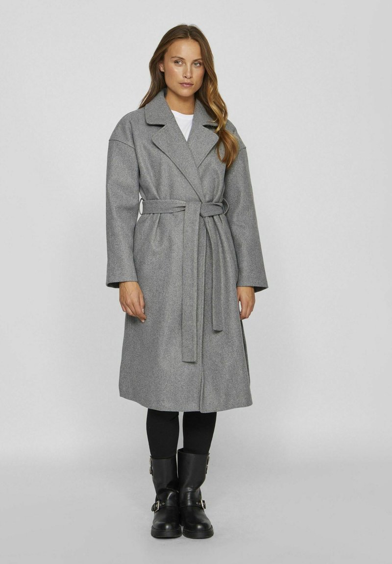 Cappotto di lana grigio con cintura, rever ampi, maniche lunghe e taglio dritto. Indossato su leggins neri e stivali alla caviglia. Texture liscia.