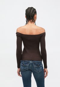 Haut marron à épaules dénudées en tissu côtelé avec manches longues, associé à un jean bleu ajusté avec poches arrière décoratives et coutures apparentes.