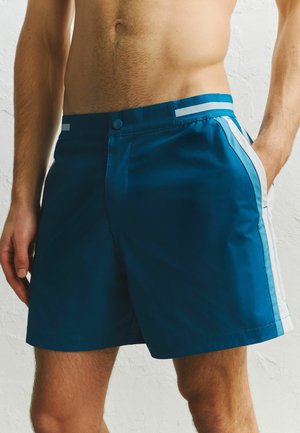 Mężczyzna w niebieskich kąpielówkach sięgających do połowy uda z jasnoniebieskimi i białymi paskami, stojący z jedną ręką w kieszeni na tle białej fakturowanej ściany.