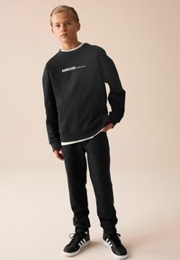 Sort sweatshirt med hvid "SUBSCRIBE" tekst, paired med matchende sorte joggers og sorte sneakers. Glat stof, afslappet pasform, ribbede manchetter.