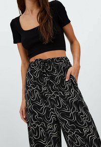 Černý žebrovaný crop top s krátkými rukávy ve spojení s černými kalhotami s vysokým pasem, zdobenými bílými abstraktními vířivými vzory a elastickým pasem.