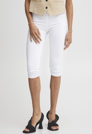 Vrouw draagt witte gerimpelde capri-broek, beige gilet met knopen en zwarte sandalen met hak en strikdetail, staand tegen een witte achtergrond.