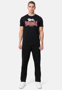 Zwarte katoenen t-shirt met rode en witte "Lonsdale London" grafische opdruk, gecombineerd met zwarte joggingbroek. Witte sneakers maken de outfit compleet.