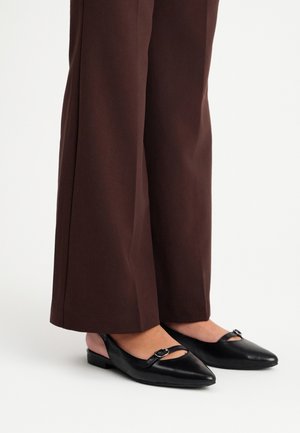 Zapatos de tacón bajo negros con punta afilada y tira con una pequeña hebilla. Material de cuero suave y suelas planas, combinados con pantalones de pierna ancha marrones.