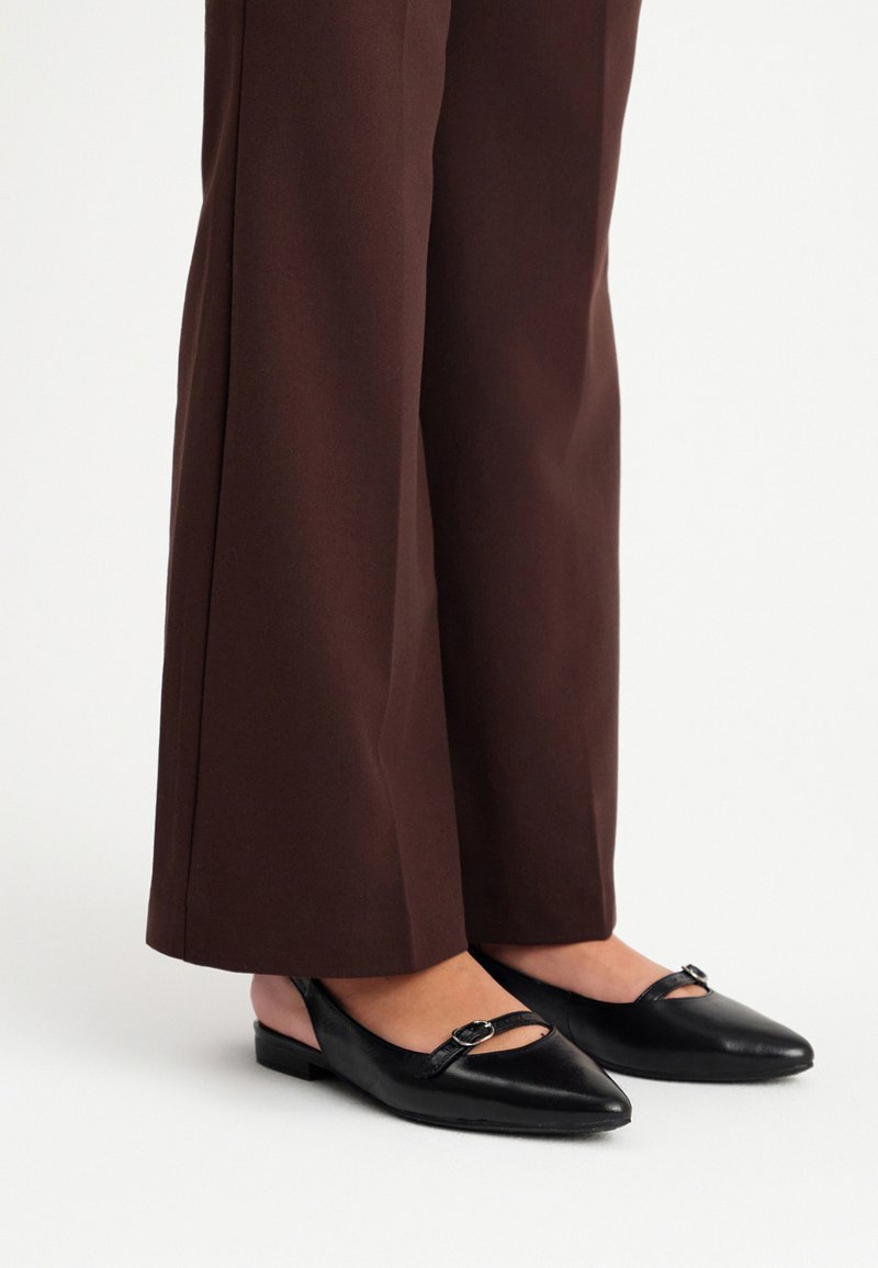 Melni ar norādītu purngalu slingback apavi ar siksniņu, kurā ir neliela sprādze. Gluda ādas materiāls un plakani zoli, kombinēti ar brūniem platām biksēm.