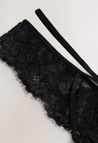 Bralette en dentelle noire avec motifs floraux et fines bretelles réglables sur un fond clair.