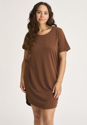 Kvinde med langt, bølget hår iført en kortærmet, knælængde brun kjole, stående foran en ensartet lys baggrund.