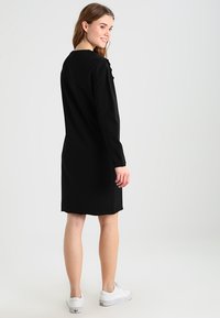 Schwarzes Langarmkleid mit geradem Schnitt, knielang, mit einer Rückennahtdetails und glatter Textur. Kombiniert mit weißen Sneakers.