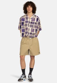 Camisa de manga corta con patrones geométricos morados sobre un fondo crema; combinada con pantalones cortos beis; usada con calcetines oscuros y zapatillas deportivas.