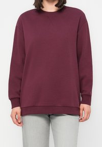 Personne portant un sweat-shirt bordeaux uni à manches longues et un pantalon gris clair, debout devant un fond blanc uni.