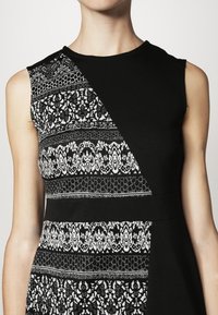 Robe noire avec un design diagonal et des motifs blancs en dentelle. Le tissu est lisse, avec une silhouette ajustée et sans manches.