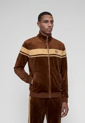 Homme portant un survêtement en velours marron avec des rayures beiges et une veste zippée, debout avec une main dans la poche, fond neutre.