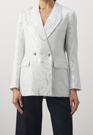 Mujer que lleva una chaqueta blanca cruzada con un sutil estampado floral y dos botones plateados, combinada con pantalones negros.